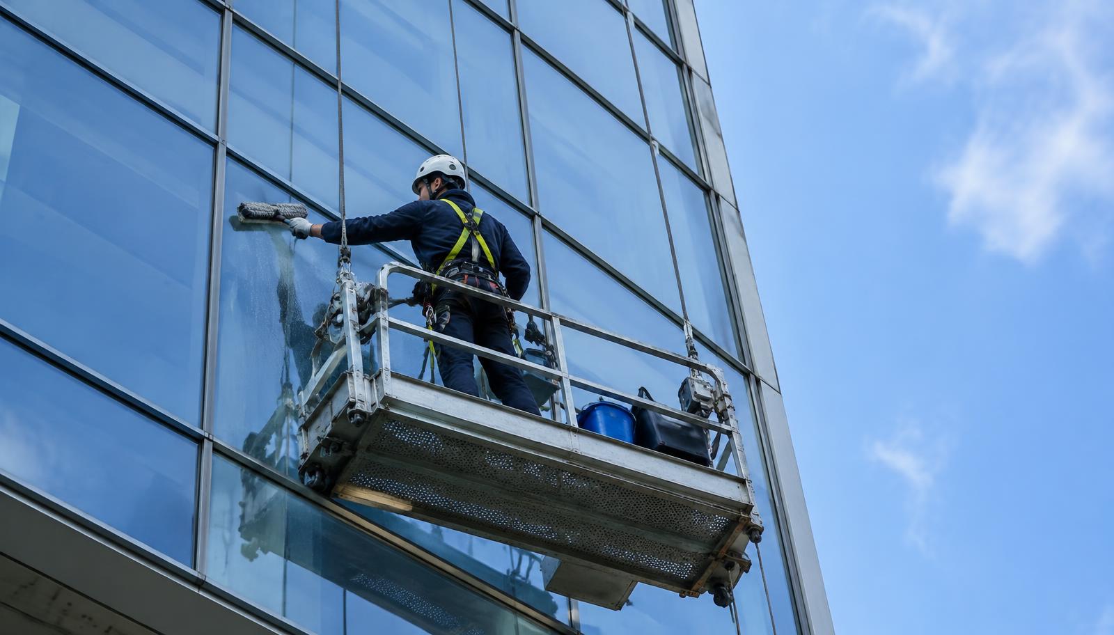 Illustrative photograph of a worker wearing a harness and helmet cleaning a building's glass facade